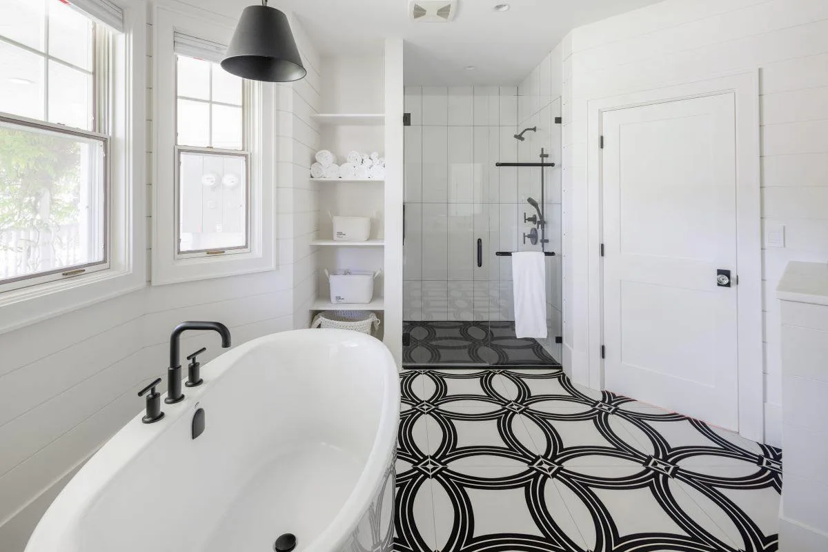 Après : salle de bains blanche avec baignoire, douche vitrée, robinetterie noire et sol à motifs.
